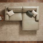 An overhead view of a light beige sectional sofa with two accent pillows, placed on a large textured beige rug over a dark wood floor, with brick walls and ceiling beams visible around the room.