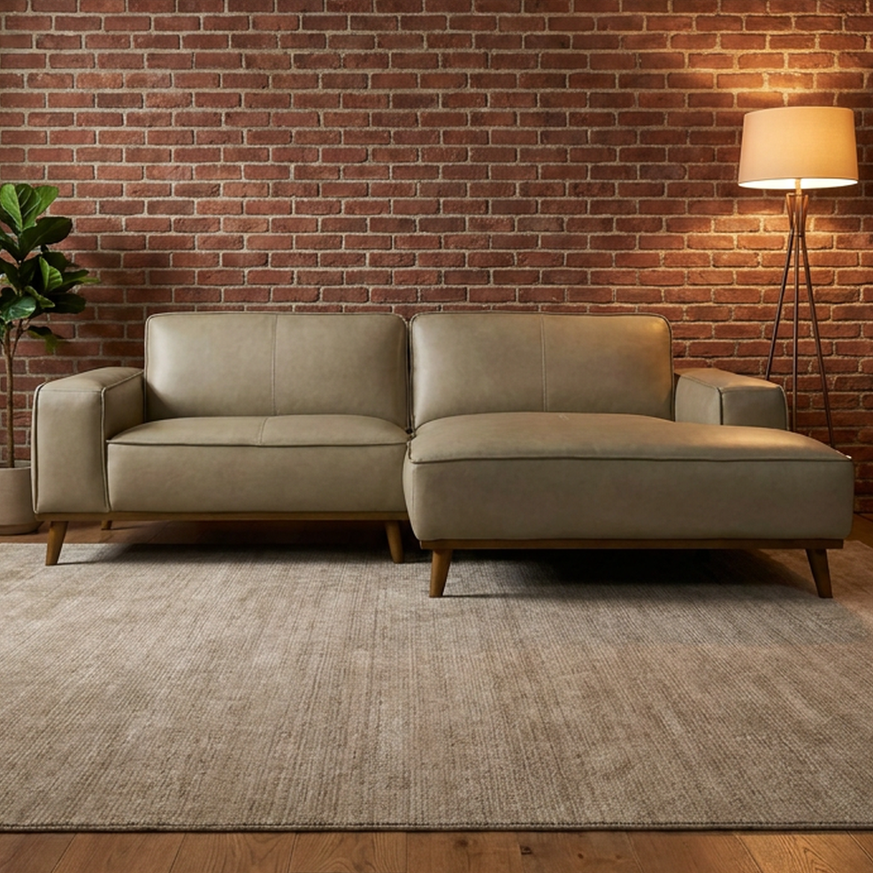A modern light beige sectional sofa with a chaise lounge, set in a living room with a brick wall, wood floor, a textured rug, a floor lamp, and a potted plant.