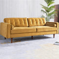 A mid-century modern three-seat sofa upholstered in mustard yellow velvet, featuring tufted back cushions, rolled armrests, and tapered dark wood legs, set against a light gray paneled wall with a potted palm in the background.