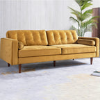 A mid-century modern three-seat sofa upholstered in mustard yellow velvet, featuring tufted back cushions, rolled armrests, and tapered dark wood legs, set against a light gray paneled wall with a potted palm in the background.