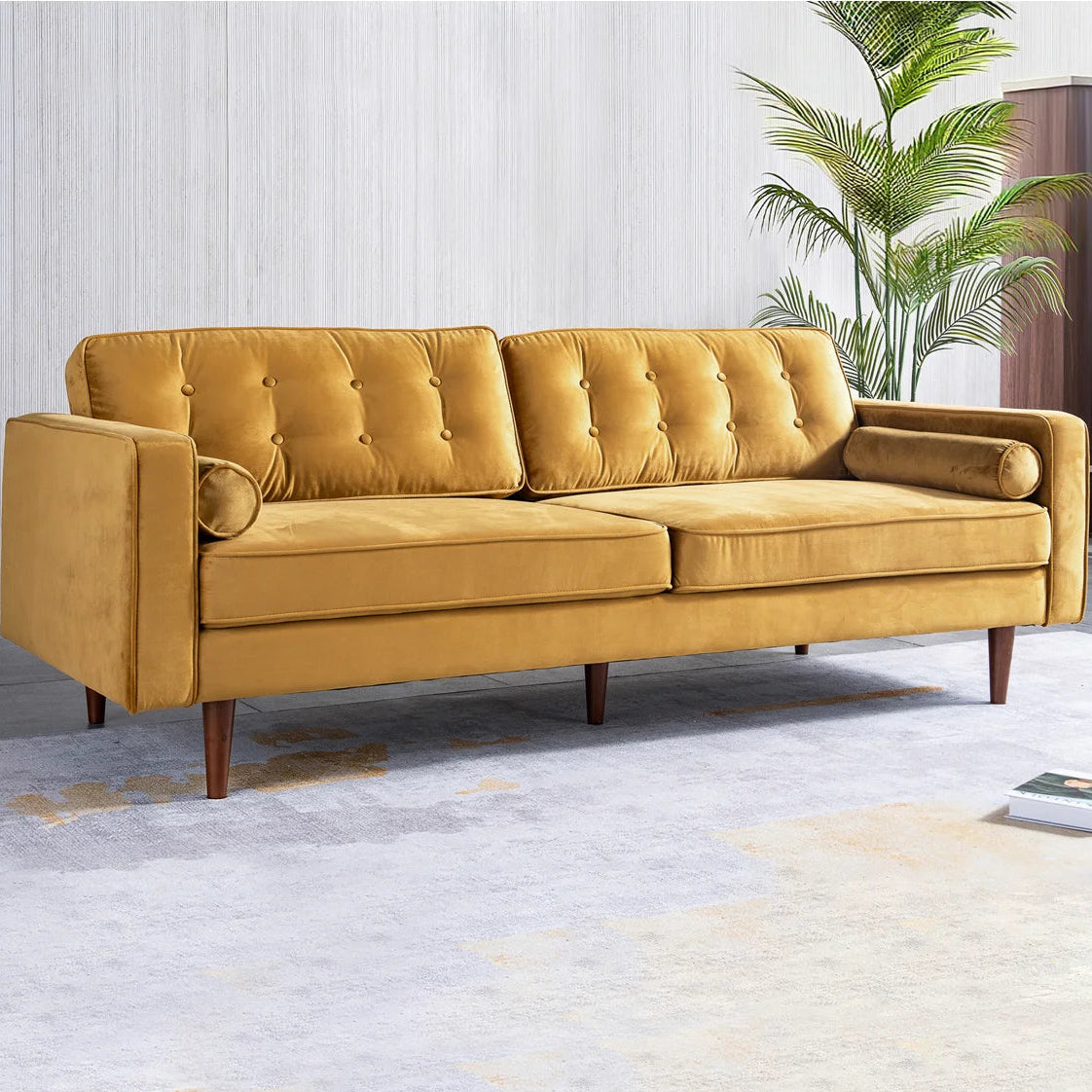 A mid-century modern three-seat sofa upholstered in mustard yellow velvet, featuring tufted back cushions, rolled armrests, and tapered dark wood legs, set against a light gray paneled wall with a potted palm in the background.