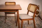 Wooden dining table with two chairs, one with a wicker back and gray cushion, on a patterned rug.