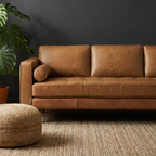 A three-seater tan leather sofa in a styled living room setting, featuring a dark charcoal wall, a potted green monstera plant, a round woven jute ottoman, and a textured beige rug.