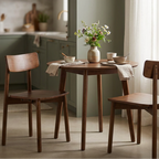 A dining set consisting of a small round wooden table and two matching chairs, styled in a bright, modern kitchen with sage green cabinets and a gold pendant light overhead.