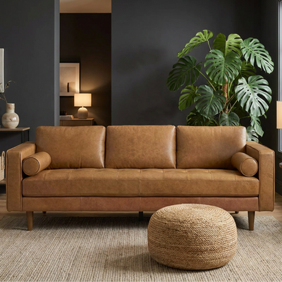 A mid-century modern three-seater sofa upholstered in warm tan leather, styled in a contemporary living room with a round woven jute pouf, dark gray walls, and a large green monstera plant.