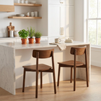 A modern kitchen scene with a white marble-topped island, two wooden bar stools, open wood shelves, and potted green plants. The room is brightly lit by a window with white cabinetry in the background.