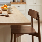 A bright, modern kitchen with cream cabinets, a light wood island, and two dark wood bar stools. On the island sits a loaf of bread on a cutting board, a small fruit bowl, and a white mug.
