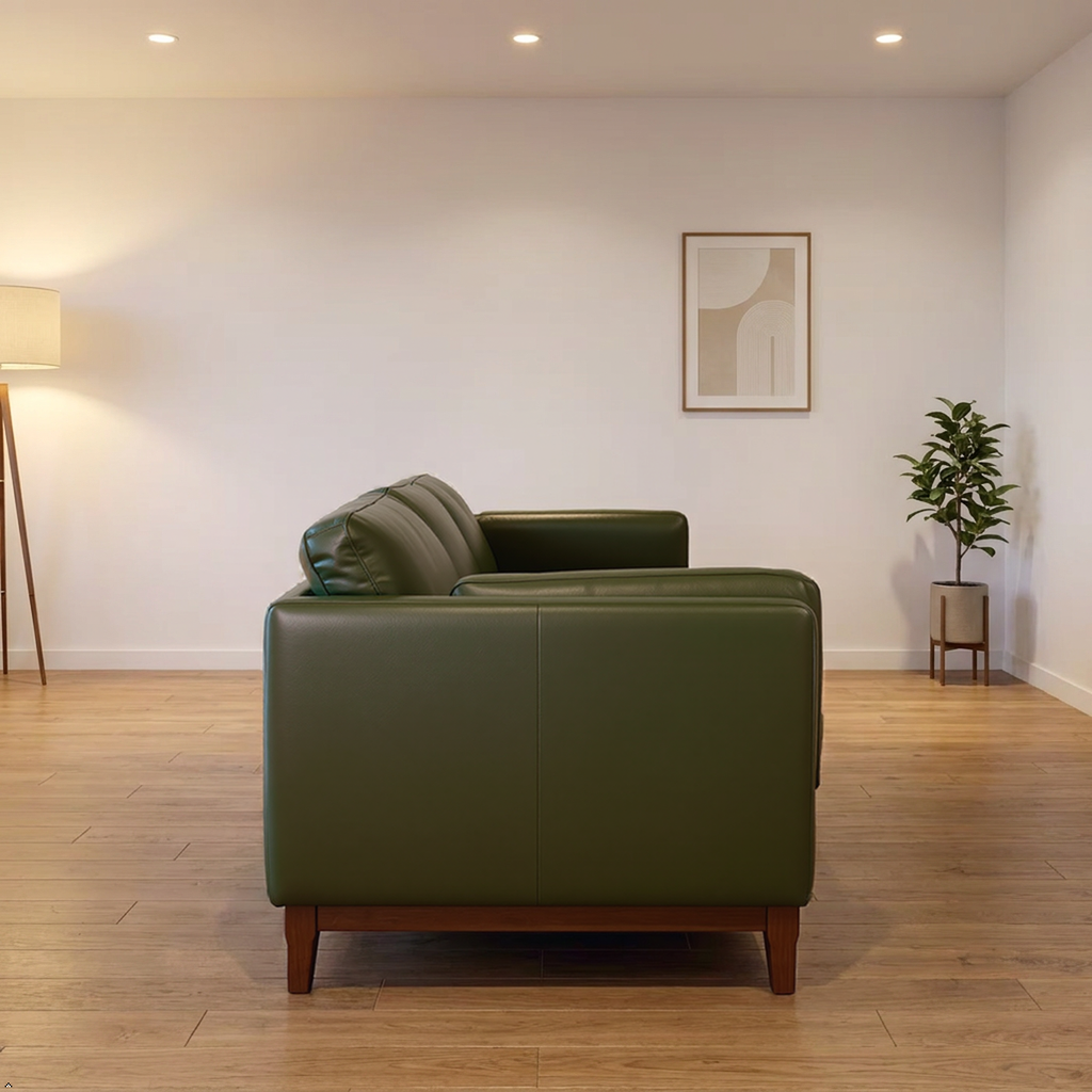 An olive green leather armchair with a dark wood base, positioned in a minimalist living room setting with light wood floors, a floor lamp, and a potted plant.