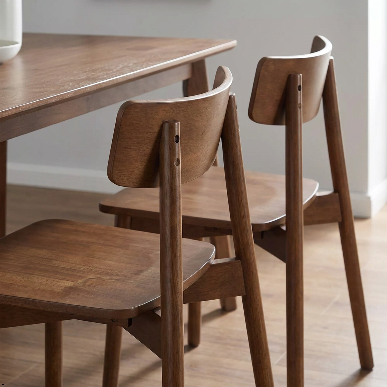 Two mid-century modern wooden dining chairs with a warm medium-brown finish, shown next to a matching wooden table in a bright room with light wood flooring.