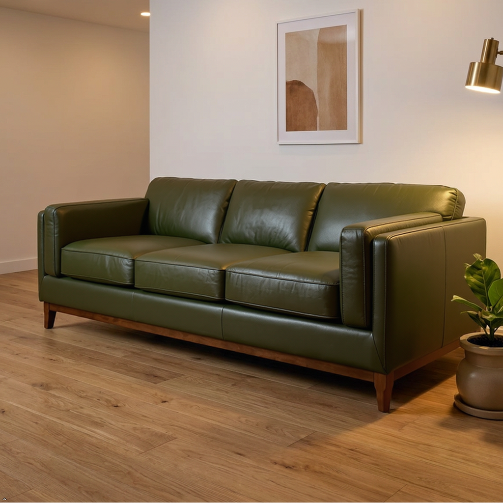 An olive green leather three-seater sofa in a modern living room setting with a wooden floor, a brass floor lamp, a potted plant, and framed wall art.