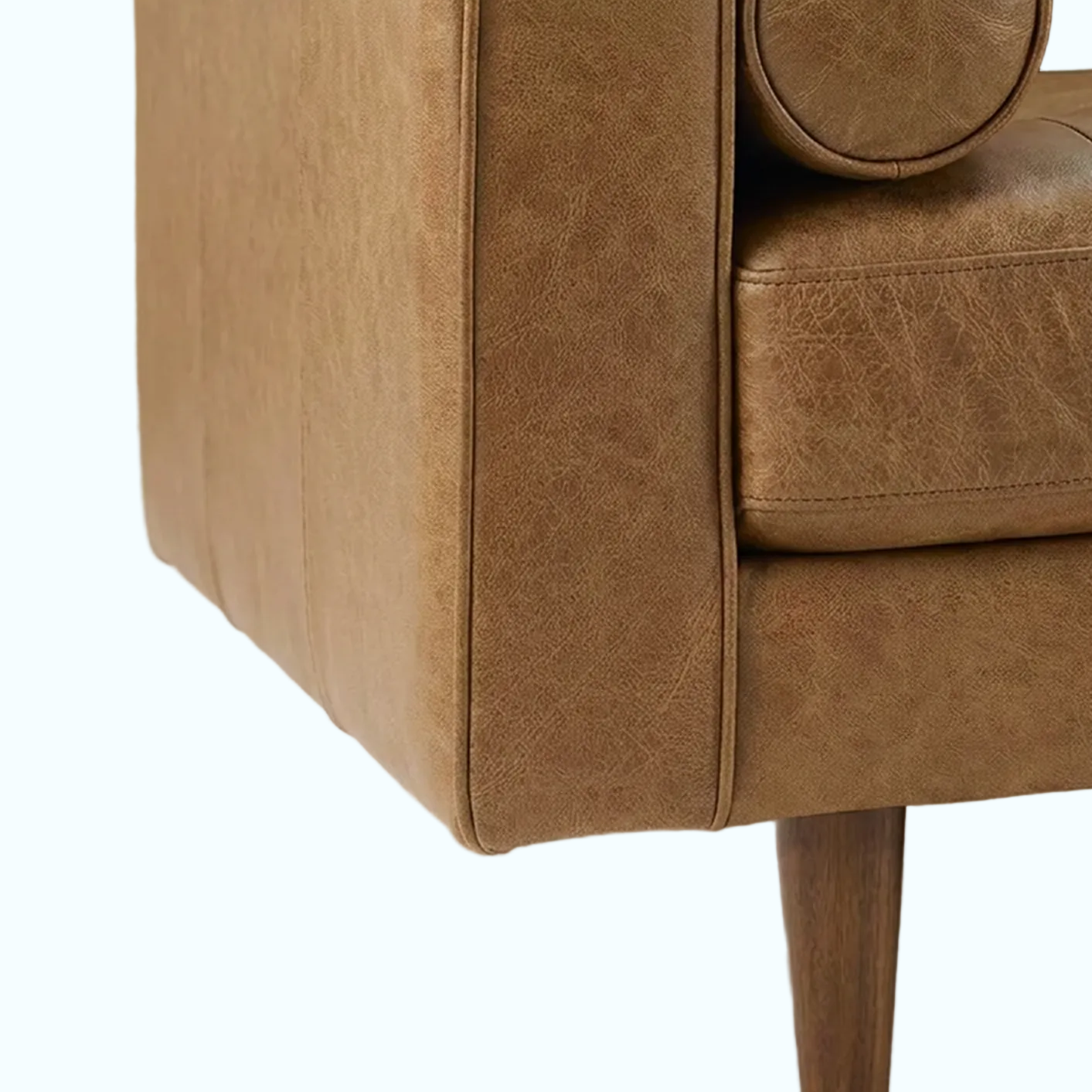 Close-up of a tan leather sofa showing the armrest, round cushion, seat, and a wooden leg on a light neutral floor.