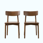 Two matching wooden dining chairs with curved backrests and angled legs, shown side by side on a light wood floor against a white wall.