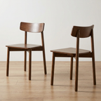 Two wooden dining chairs with a warm medium-brown finish, shown from different angles on a light wood floor against a plain off-white wall.