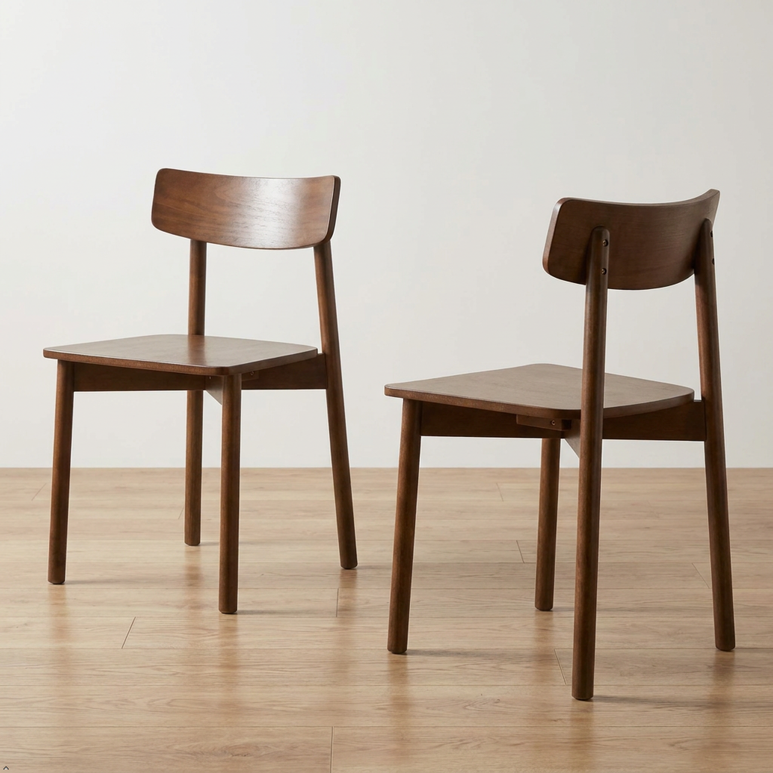 Two wooden dining chairs with a warm medium-brown finish, shown from different angles on a light wood floor against a plain off-white wall.
