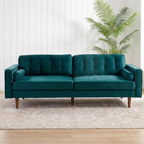 A teal green velvet loveseat sofa with tufted back cushions and tapered wooden legs, positioned on a light beige rug in a bright room with white paneled walls, a potted palm, and windows.