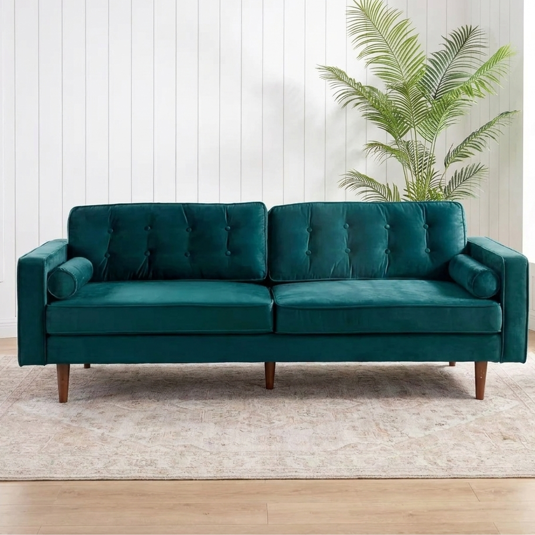 A teal green velvet loveseat sofa with tufted back cushions and tapered wooden legs, positioned on a light beige rug in a bright room with white paneled walls, a potted palm, and windows.