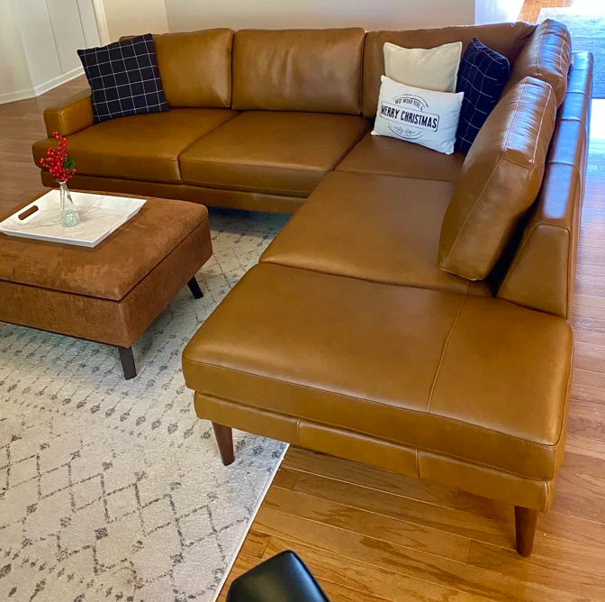 A tan leather sectional sofa with a chaise lounge in a living room setting, styled with accent pillows and a matching brown leather ottoman on a patterned rug.